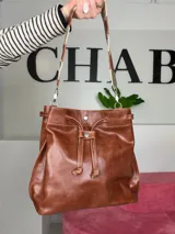 Bolso tipo bucket bag Chabas modelo 3.1, confeccionado en cuero genuino de color marrón. Cuenta con cierre de cordón ajustable, dos tipos de asas (una corta de cuero con detalles decorativos y otra larga tipo bandolera con tira textil) y diseño sin forro interior.