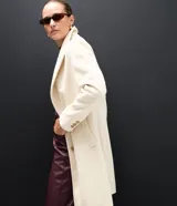 Saco femenino de corte alargado, color crema, con solapa amplia, cierre frontal de dos botones y bolsillos laterales embutidos.