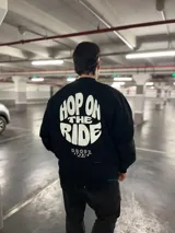 Buzo negro de algodón con interior suave y fit relajado. Presenta un estampado grande en la espalda con el texto "Hop On The Ride" en tipografía bold blanca y un diseño circular, debajo del cual se encuentra el logo "Dropz Studio Est. 2025".