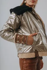 Campera puffer metalizada en tono dorado, con interior y detalles en corderito beige. Presenta capucha negra de corderito, cierre frontal y puños ajustables con tiras de cuero y tachas metálicas.