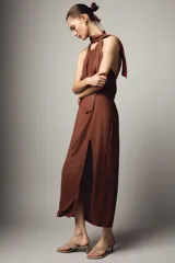 Vestido midi de color marrón con cuello halter y lazada en la nuca. Presenta un diseño con espalda descubierta, frunces en la cintura y una abertura lateral en la falda.