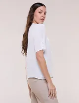 Blusa blanca de corte recto y tejido liviano de gasa con forro interno delantero. Presenta escote en V con cuello redondo y lazo central para anudar, mangas cortas con hilera de botones forrados en los laterales y ruedo ligeramente redondeado.