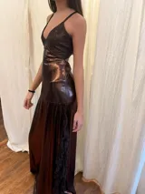 Vestido largo marrón metalizado con corte bajo el busto, escote en V, tirantes finos cruzados en la espalda y aberturas cut-out laterales en la cintura. Presenta falda larga con tajo lateral adornado con volados de encaje.
