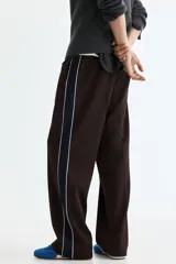 Pantalón deportivo marrón de punto con leve brillo, corte relajado y piernas rectas con pliegues cosidos. Cuenta con franjas laterales oscuras y ribetes blancos contrastantes, cintura elástica y bolsillos laterales.