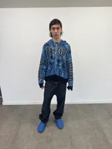 Buzo tipo hoodie tejido de punto grueso con cierre frontal completo hasta la capucha. Presenta un diseño gráfico abstracto en tonos azules y negros, con tipografía estilo gótico en el frente y las mangas. Cuenta con puños y ruedo acanalados en color negro, y etiqueta de marca en la manga.