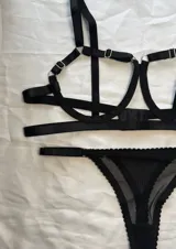 Conjunto de lencería negro de tiras elásticas con arandelas metálicas, compuesto por un soutien tipo arnés y una bombacha de encaje y malla transparente.
