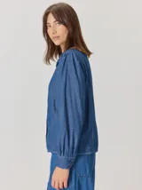 Camisa de denim con silueta levemente entallada, cuello bebé y mangas largas con sutil frunce en los hombros. Presenta costuras curvas decorativas con pespuntes en contraste y cierre frontal con botones.