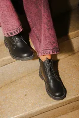 Bota tipo borcego de cuero negro, con cordones frontales, cierre lateral y suela track con costura blanca visible.