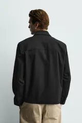 Sobrecamisa negra de corte relajado, confeccionada en tejido con mezcla de algodón elástico. Presenta cuello solapa, cierre frontal de cremallera, bolsillos de plastrón en el pecho y bolsillos de vivo en la cadera.