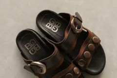 Sandalias bajas de cuero marrón con doble tira, hebilla ajustable y apliques de tachas metálicas.