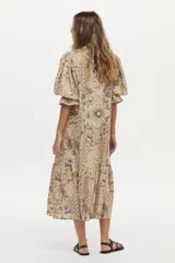 Vestido túnica de lino y viscosa, color camel con estampa artesanal en tonos oscuros, escote en V profundo, mangas amplias abullonadas y falda larga con volado en el ruedo.