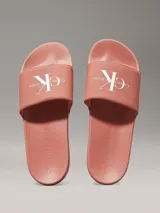 Ojotas tipo slide de color rosado, con base de EVA y correa ancha de lona de algodón reciclado. La correa presenta el logo de Calvin Klein Jeans bordado en blanco.