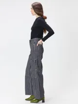 Pantalón de tiro medio y pierna ancha confeccionado en denim de algodón grueso. Presenta un diseño a rayas verticales en tonos azul y celeste, con costuras en contraste color rojo, bolsillos laterales tipo cargo y remaches metálicos.