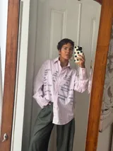 Camisa oversize de corte recto, confeccionada en tela de algodón a rayas verticales celestes y blancas. Presenta cuello clásico, cierre frontal con botones y mangas largas. El diseño destaca por una estampa frontal con palabras relacionadas al amor en diversos idiomas en color bordó.