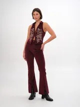 Chaleco de pana color bordó, ajustado al cuerpo, con pinzas y elásticos en la espalda baja. Presenta un largo irregular, cuello clásico camisero y cierre frontal con ganchos peleteros. Tiene bordados decorativos en color crema en el frente.