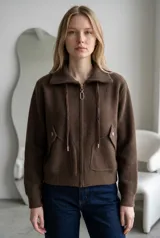 Campera de paño color marrón, con cuello alto, cierre frontal completo y cordones decorativos. Presenta bolsillos frontales con solapa y cierre a presión, y puños tejidos.