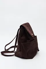 Mochila de símil gamuza color negro con textura tipo reptil. Presenta un diseño minimalista con solapa frontal, cierre de cordón ajustable y bolsillos laterales. Incluye un detalle metálico rectangular en la solapa.