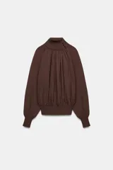 Blusa marrón de punto fluido, con cuello subido abotonado en un lateral y mangas largas con puños ajustados. Presenta un diseño fruncido en la parte frontal que aporta volumen.