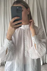 Camisa blanca de estilo romántico con textura de plumeti y rayas verticales. Presenta cuello alto con volados, cierre frontal con botones forrados y mangas largas abullonadas con puños ajustados.