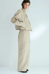 Campera corta de sarga de algodón, color beige, con cuello elevado ajustable por cordón, solapa rompevientos con botones a presión ocultos y cierre. Presenta bolsillos delanteros insertados, mangas ranglán con hombros caídos y bajo globo que genera una silueta abullonada.
