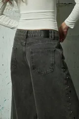 Jean de corte wide leg en color gris lavado, con diseño de aberturas laterales decoradas con tachas metálicas a lo largo de toda la pierna.