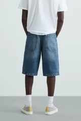 Bermuda de denim de algodón, color azul medio lavado, de corte ancho (baggy fit) y largo hasta la rodilla. Presenta cinco bolsillos y cierre frontal con cremallera y botón.