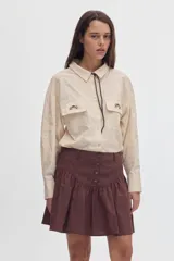 Camisa de tacto de seda color beige, entallada en la cintura, con cuello camisero, bolsillos frontales con solapa y bordados, y cordón decorativo en el cuello.