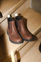 Bota tipo Chelsea de cuero marrón con textura granulada, paneles laterales elásticos en color negro y suela track dentada con costura blanca visible.