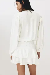 Vestido corto de color blanco en tejido con textura, con escote redondo, abertura con tiras para anudar, mangas largas abullonadas y falda de volados escalonados.