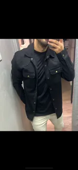 Sobrecamisa de gabardina color negro, con cierre frontal de botones metálicos a presión, bolsillos frontales con solapa y mangas largas.
