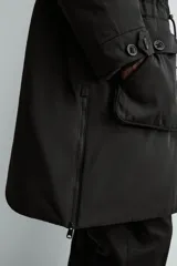 Parka técnica de corte relajado con acabado repelente al agua. Presenta cuello subido con capucha ajustable, cierre frontal con cremallera oculta por solapa con botones y cintura ajustable con cordón. Incluye múltiples bolsillos frontales con solapa, un bolsillo con cremallera en la manga y puños con interior en rib. El diseño cuenta con interior acolchado y aberturas laterales con cremallera en el bajo.