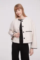 Chaqueta corta de paño color marfil, con cuello redondo, cierre frontal de botones negros y dos bolsillos frontales. Presenta detalles de costuras decorativas en contraste negro en los bordes de los bolsillos, puños y ruedo.