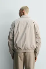 Cazadora regular fit de tejido técnico color beige, con cuello subido, cierre de botones y cremallera frontal, bolsillos de doble vivo en la cadera y acabados elásticos.