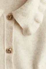 Saco corto de punto suave color beige, con cuello camisero con ribete de volados y botones de metal con diseño de nudo. Tiene hombros ligeramente caídos y remate acanalado en puños y bajo.