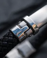 Pulsera de cuero trenzado negro con placa central de acero inoxidable y cierre de seguridad metálico.