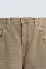 Jeans de hombre, tiro medio, color marrón claro o caqui, con corte relaxed fit y perneras anchas. Presenta cinco bolsillos y costuras laterales giradas hacia el frente.