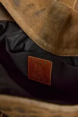 Cartera de ante color marrón con terminación gastada, cuerpo blando, doble asa de mano, cierre frontal con hebilla y detalles de tachas. Incluye cordones de ajuste en los laterales y un colgante metálico decorativo.