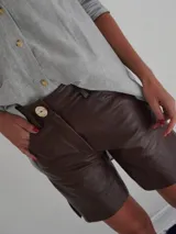 Bermuda de cuero color marrón oscuro con efecto desgastado, tiro medio, cierre frontal con botón y bolsillos.