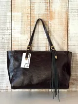 Cartera tipo tote de cuero marrón oscuro con acabado brillante y efecto envejecido. Presenta doble asa con hebillas metálicas decorativas y un detalle de flecos largos colgantes en uno de los laterales.