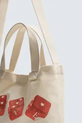 Bolso tipo shopper de tela color beige, con asas cortas y asa de hombro. Estampado frontal con cuatro dados rojos con corazones y texto que dice "SOMETIMES SÍ, SOMETIMES BYE".