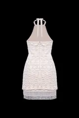 Vestido corto de crochet color beige con escote halter y espalda nadadora. Presenta inserciones de malla transparente en la cintura, bordado de perlas y cristales, y un ruedo con flecos largos de cuentas.