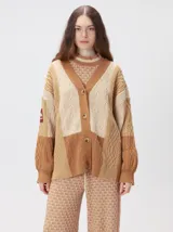 Cardigan oversize de tejido de punto grueso con diseño de bloques de color en tonos beige y marrón. Presenta cuello en V, cierre frontal con botones, mangas largas abullonadas y parches decorativos en el pecho y el hombro.