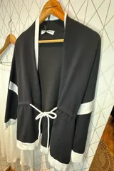 Cárdigan de punto en color gris con detalles en negro en los bordes, puños y una franja horizontal en las mangas. Presenta un diseño abierto con cordón ajustable en la cintura y bolsillos laterales.