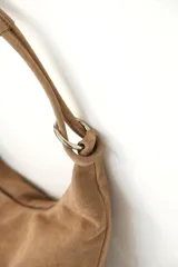 Cartera tipo hobo de gamuza color marrón, con diseño semicircular, asa de hombro y argolla metálica decorativa en la unión del asa.