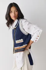 Chaleco de lana color marrón con cuello estilo bebé, cierre frontal con botones grandes y detalle de puntilla en cuello, sisas y bolsillo frontal. Presenta una franja decorativa en color rojo en el ruedo.