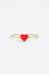 Pulsera rígida tipo esclava confeccionada en latón dorado con un dije central en forma de corazón esmaltado en color rojo, que incluye el nombre de la marca grabado en el frente.