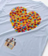 Remera boxy de algodón color celeste, con cuello redondo y mangas cortas. Presenta un estampado frontal con lettering y un estampado trasero grande con un corazón formado por círculos coloridos y una pequeña ilustración de una casa debajo, inspirado en la película "Up: Altas Aventuras".