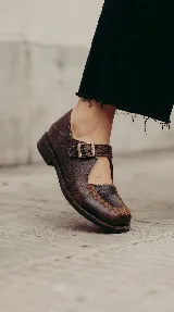 Mocasín de cuero marrón con acabado texturizado, diseño tipo Mary Jane con tira ajustable mediante hebilla lateral y detalle de costuras artesanales visibles en la puntera.