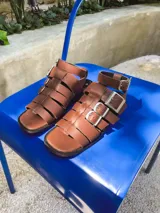 Sandalias planas de cuero color marrón, con tiras anchas y hebillas plateadas.