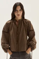 Blusa de color marrón con estampado de rayas verticales y pequeñas flores blancas. Presenta cuello redondo con lazo de ajuste, cierre frontal con botones y mangas largas abullonadas con detalles de encaje calado y puños ajustables con lazo.